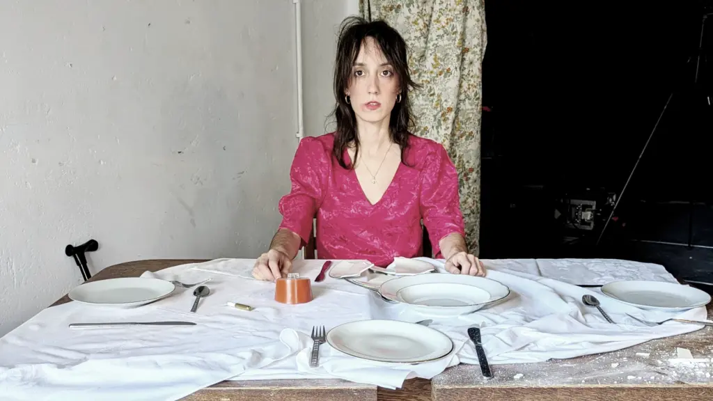Performance-Künstlerin Mariana Nicotra sitzt an einem schäbigen Esstisch mit zerstörten Tellern und starrt ausdruckslos ins Leere